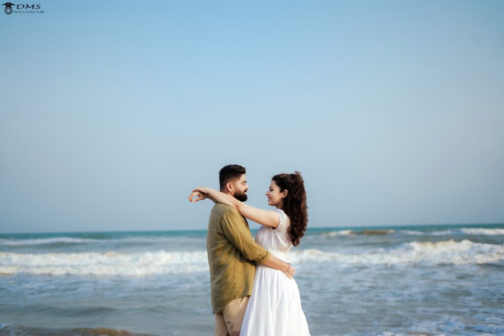 A couple is standing T the beach & looking together with their hands on each other shoulder and having a background of ocean.