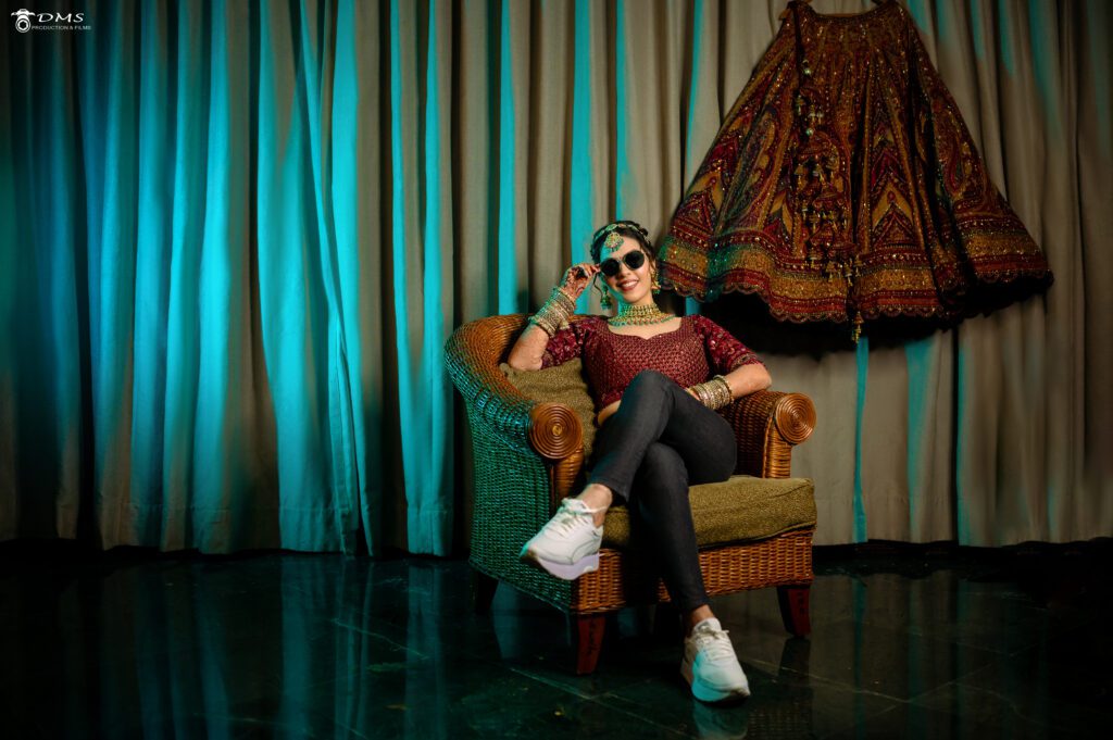 a wedding bride with a sunglasses sitting in a sofa chair with one leg on other leg, poses for wedding photography and in background bride lehnga is hanging