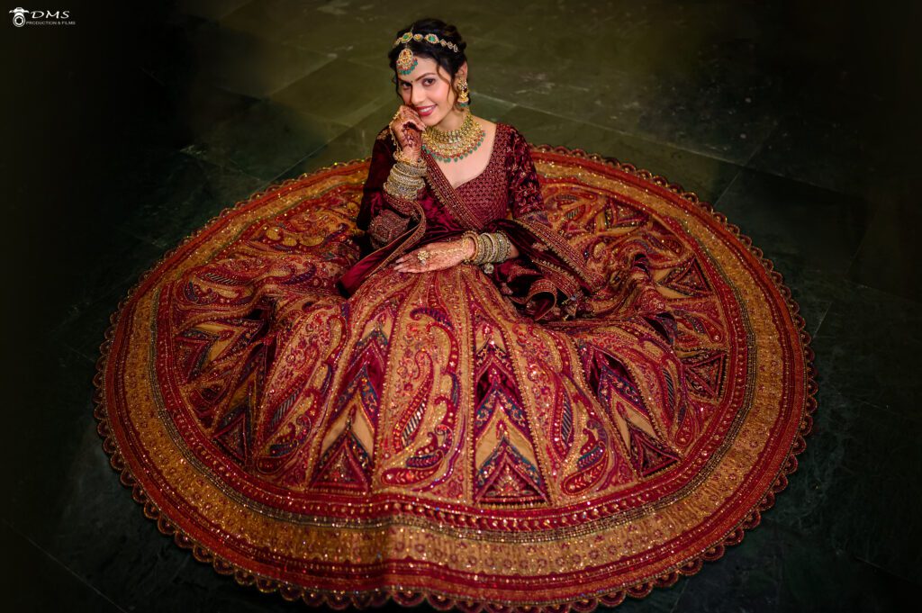 A radiant Indian bride in a maroon lehenga with ornate gold embroidery, perched gracefully on the floor, gazes up with joy and anticipation on her wedding day